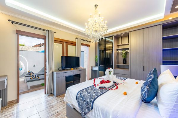 Luxurious Bali villa bedroom with king bed adorned in rose petals and towel swan, chandelier lighting, TV, wardrobes, and open doors to private pool courtyard.